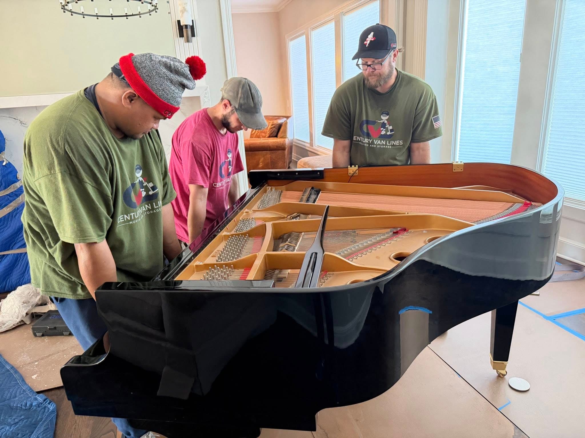 Three professional movers examine the internal strings of a black grand piano inside a home.