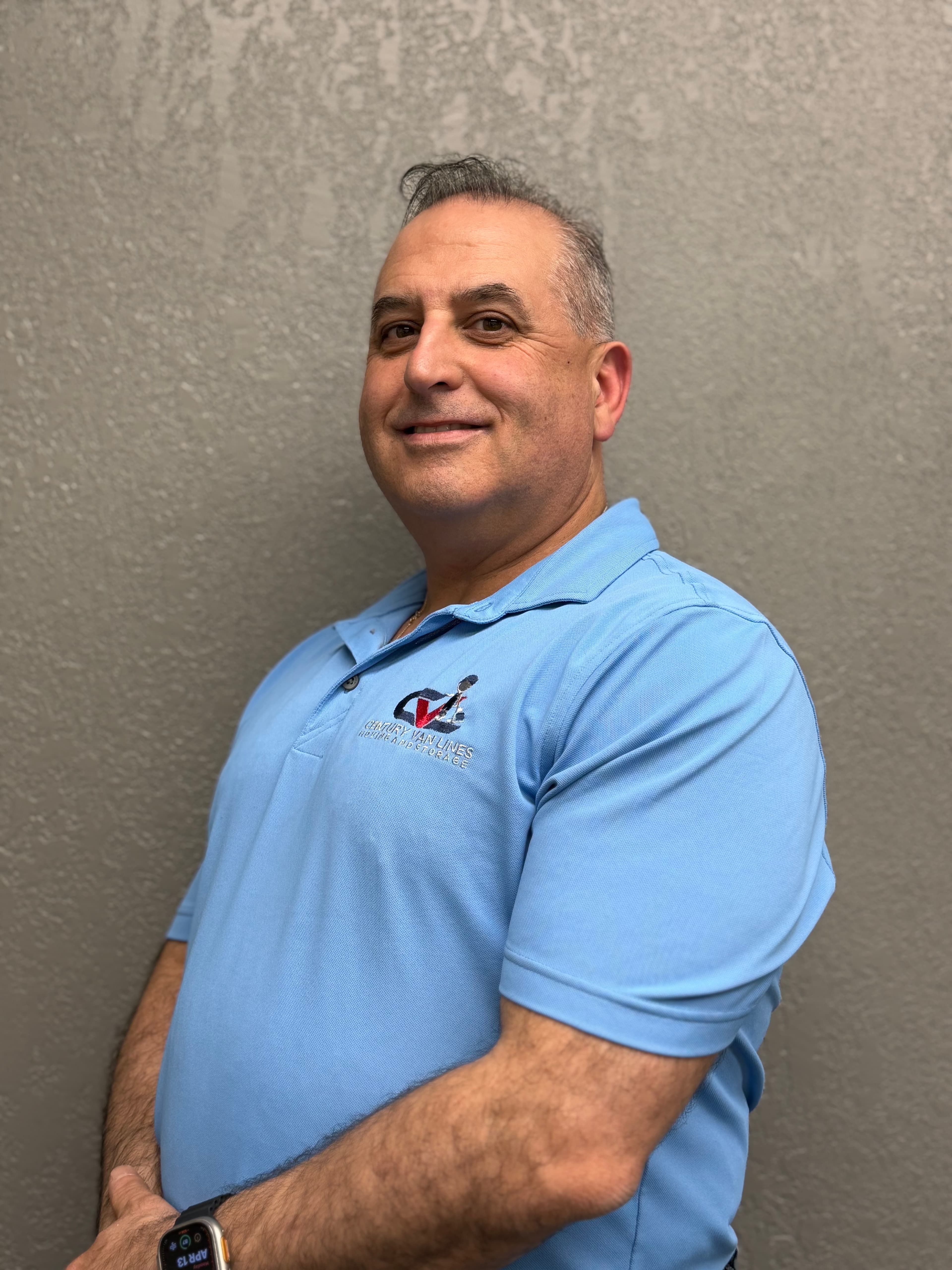 Smiling man wearing a light blue polo shirt with a logo against a textured grey wall.