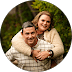 Smiling woman hugging a man from behind outdoors, set against a backdrop of green foliage.